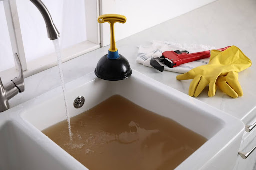 Plumbing tools and gloves on a countertop near a clogged sink full of dirty water.